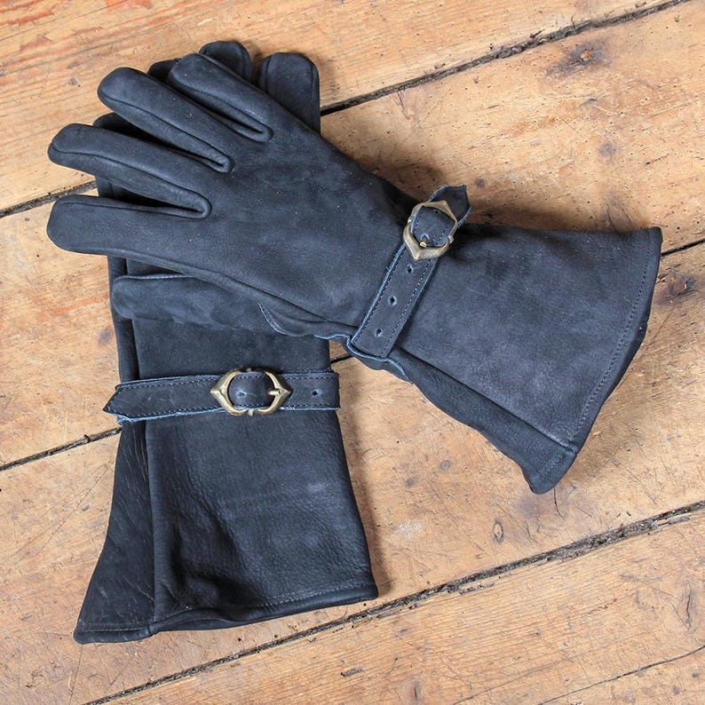 May include: A pair of black leather gloves with brass buckles on the wrist straps. The gloves are lying on a wooden surface. The gloves appear to be new and unworn.
