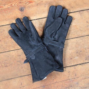 May include: A pair of dark gray leather gloves with long cuffs and buckle details. The gloves are made of a soft, textured leather and are laid out on a wooden surface. The gloves appear to be new and unworn.