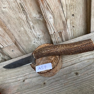 Peut inclure: Un couteau à lame fixe doté d'une lame en métal sombre et texturé, avec un manche en bois de cerf brun et rugueux. Le couteau repose sur une base en bois circulaire, portant une petite étiquette blanche avec le texte AS4 écrit en bleu.