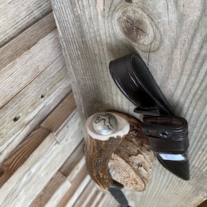 Peut inclure: Un couteau de chasse avec un manche en bois de cerf brun texturé, surmonté d'un capuchon en métal argenté orné d'un emblème de cerf en relief. Il est accompagné d'un étui en cuir marron foncé doté d'un passant et d'une étiquette blanche.