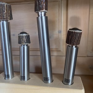 Peut inclure: Quatre bouchons de bouteille cylindriques en métal de hauteurs variées, chacun surmonté d'un capuchon brun texturé rappelant un bois de cerf. Les corps en métal argenté portent le texte noir "BRASS WIESN FESTIVAL" imprimé près de la base.