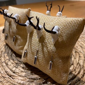 Könnte beinhalten: Zwei Kissen aus Jute mit dekorativen Hirschgeweih-Akzenten. Jedes Kissen hat kleine, silberfarbene Metallstifte und künstliche Hirschgeweihe. Die Kissen liegen auf einer gewebten, kreisförmigen Matte.