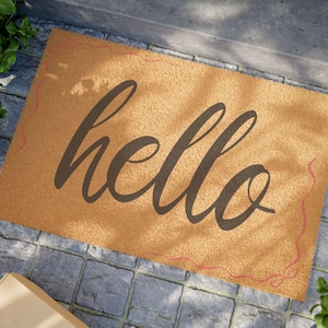 Puede incluir: Una alfombra de puerta rectangular hecha de fibra de coco de color beige. La palabra "hello" está escrita en una escritura marrón oscuro en el centro. Un borde decorativo rosa fino con un diseño de remolino enmarca los bordes de la alfombra.