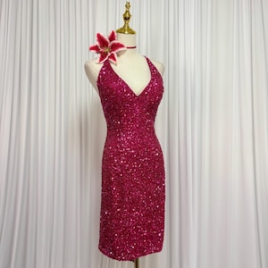 May include: A magenta sequined halter dress on a mannequin. The dress has a V-neck and a fitted silhouette. A red and white flower and a matching choker adorn the mannequin's neck. The background is a white curtain.