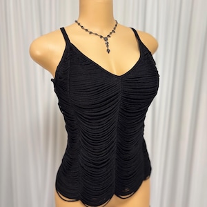 May include: Black tank top with a V-neck and spaghetti straps. The top has a ruched design and a scalloped hem. A dark necklace with a pendant is also visible.