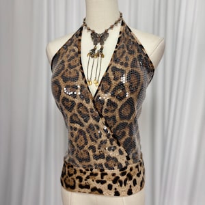 May include: A halter top with a leopard print design. The top is brown and tan with black spots and is covered in sequins. A butterfly pendant necklace with gold-colored charms is draped over the top.