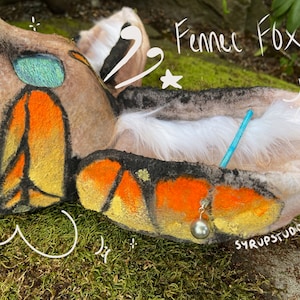 Puede incluir: Primer plano de un juguete de peluche Fennec Fox con pelaje naranja y amarillo. El juguete está acostado en un lecho de musgo verde. El juguete tiene una cola blanca y un ojo azul y verde. El texto "Fennec Fox" está escrito en blanco sobre un fondo verde. El texto "SYRUPSTUDIOS" está escrito en blanco sobre un fondo marrón.