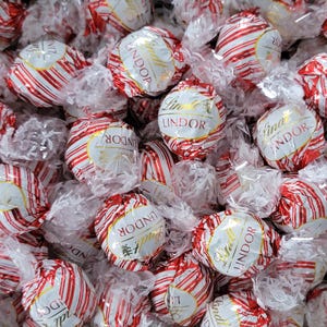 May include: A close-up shot of numerous individually wrapped Lindor chocolate truffles. The wrappers are red and white striped with gold accents and the Lindor logo. The candies are partially visible through the clear cellophane.