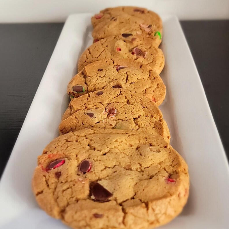 Puede incluir: Cinco galletas reci&eacute;n horneadas dispuestas en un plato rectangular blanco. Las galletas, de color marr&oacute;n dorado, est&aacute;n salpicadas de chispas de chocolate y caramelos de colores. La imagen destaca la textura y los ingredientes.