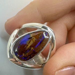 May include: A silver ring featuring a large, oval opal gemstone. The opal displays a vibrant array of colors, including blues, purples, and golds. The ring's design has a modern, openwork style, with the gemstone securely set within the silver band.