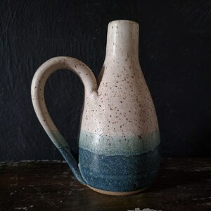 Puede incluir: Una jarra de cerámica blanca con un esmalte moteado y una banda de esmalte azul y verde. La jarra tiene un asa y una abertura estrecha.