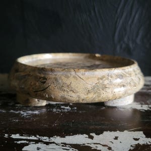 May include: A round, beige stone bowl with a textured surface and three small feet. The bowl has a wide, shallow design and is likely used for decorative purposes.