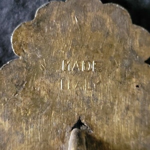 May include: A gold-colored metal pin with a flower design. The back of the pin has the words "MADE IN ITALY" engraved on it.