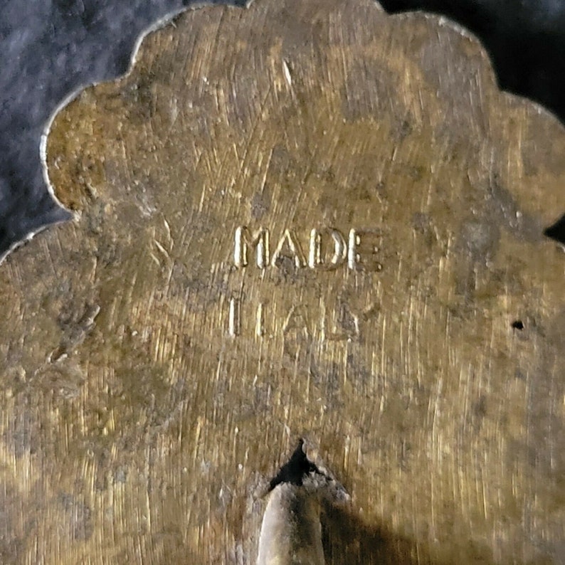 May include: A gold-colored metal pin with a flower design. The back of the pin has the words "MADE IN ITALY" engraved on it.