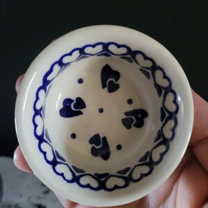 Könnte beinhalten: Kleine, runde, cremefarbene Keramikschale mit blauem Herzmuster. Der Rand ist mit einer Kette aus blauen Herzen verziert. Das Innere zeigt verstreute blaue Herzformen und Punkte. Ein dekoratives Stück.