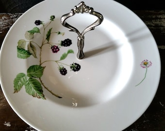Könnte beinhalten: Weiße Keramik-Servierplatte mit silbernem Griff. Die Platte hat ein Design aus schwarzen und grünen Brombeerranken mit einer einzelnen rosa Margerite an der Seite.