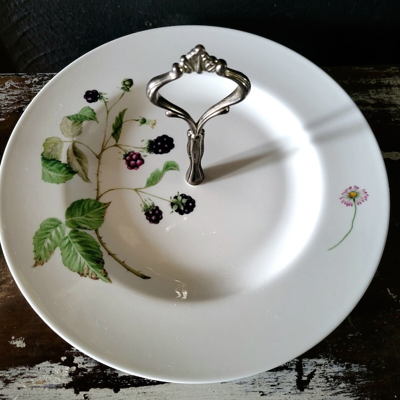 May include: White ceramic serving plate with a silver handle. The plate has a black and green blackberry vine design with a single pink daisy on the side.