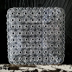 Könnte beinhalten: Ein quadratischer Teller aus klarem Glas mit einem sich wiederholenden geometrischen Muster aus gepressten Sternen und kleinen kreisförmigen Vertiefungen. Der Teller hat einen gewellten Rand und eine strukturierte Oberfläche.