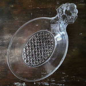 Puede incluir: Fuente de vidrio transparente con forma ovalada y asa decorativa. El centro de la fuente presenta un diseño texturizado en forma de diamante. El asa tiene un diseño floral y de hojas.