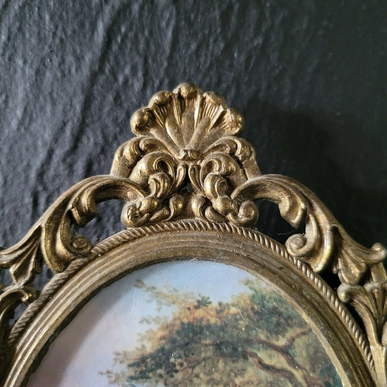 May include: A gold-toned ornate picture frame with a detailed floral design. The frame has a glass oval insert and is perfect for displaying a small photograph or artwork.