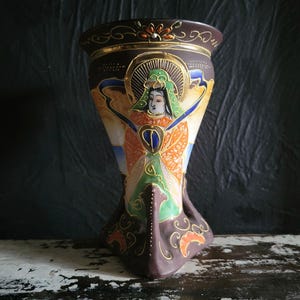 May include: A brown ceramic vase with a flared top and a detailed painted design. The vase features a central figure with a halo, wearing a green headdress and orange robes. Gold accents and floral patterns adorn the rim and base. The vase is approximately 10 inches tall.