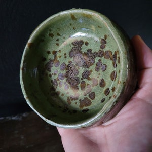 May include: A green ceramic bowl with a speckled brown glaze. The bowl has a rounded shape and a slightly flared rim.