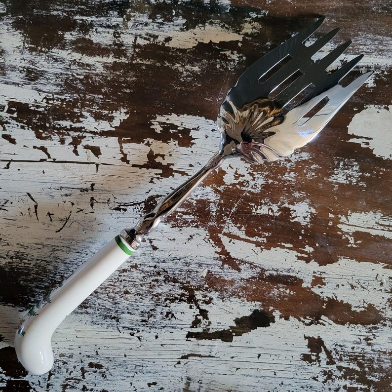 May include: A silver serving fork with a white handle. The handle features a green band near the fork head and floral details. The fork tines are broad and flat. The handle is curved for comfortable use.