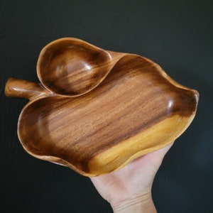 May include: A wooden serving dish shaped like a leaf with a smaller bowl carved into the side. The dish is made of dark brown wood with a natural grain pattern.