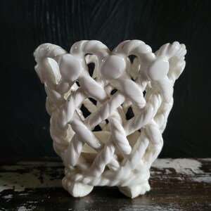 Peut inclure: Un récipient en céramique blanche présentant un design complexe en treillis ajouré qui ressemble à une corde tressée. La structure est cylindrique avec un bord supérieur évasé et repose sur une base formée par le motif en céramique tissée.