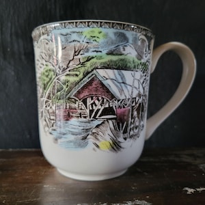 May include: A white ceramic mug featuring a colourful illustration of a covered bridge over a stream. The scene includes green trees, a mountain in the background, and a red-roofed structure. The mug has a curved handle and a decorative border pattern.