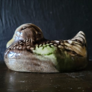 May include: A ceramic duck figurine with a brown, green, and white speckled glaze. The duck is in a swimming position with its head turned to the side.