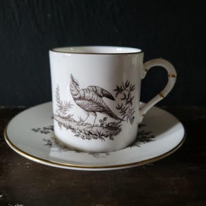May include: A white porcelain teacup and saucer set with a black and white bird design. The cup has a gold rim and a gold band around the handle. The saucer also has a gold rim.