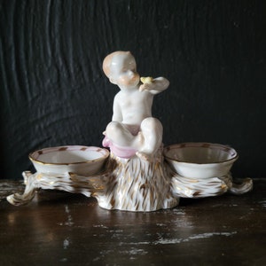 Puede incluir: Figura de porcelana que representa un querubín sentado sosteniendo un pequeño objeto amarillo. El querubín está sentado sobre un tocón de árbol con dos cuencos pequeños a cada lado. Los cuencos y el tocón son blancos con detalles dorados y ribetes rosas.