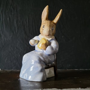 May include: A porcelain figurine of a rabbit wearing a blue nightgown and holding a cup and a piece of yellow cheese. The rabbit is sitting on a wooden chair.