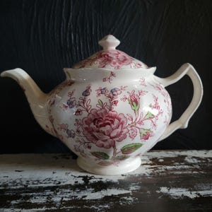 May include: A white ceramic teapot with a lid, featuring a floral pattern in shades of pink, green, and purple. The teapot has a curved spout and a handle, and sits on a white base. The background is a dark, textured surface.
