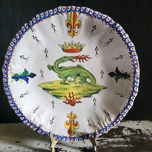 May include: White ceramic plate with a hand-painted design. The plate features a green dragon breathing fire, a crown, and fleur-de-lis symbols. The rim has a blue and red scalloped border. Decorative plate.
