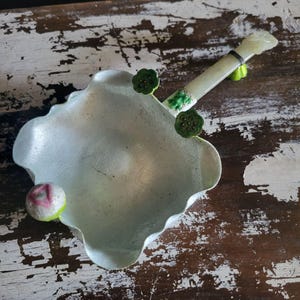 May include: A light green, lotus-shaped dish with a matching handle. The dish has a textured surface and is decorated with small green lotus flower accents. A small, round, pink and white accent is on the side of the dish. The handle is made of a light green stone.