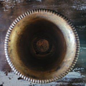 May include: A gold metal vase with a decorative rim. The vase is empty and has a worn, aged look.