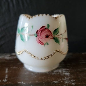 May include: A white, egg-shaped glass cup with a scalloped rim and a gold trim. The cup is decorated with a hand-painted pink rose and green leaves. The base of the cup has a small foot.