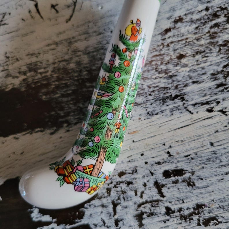 May include: A white ceramic Christmas stocking featuring a detailed illustration of a decorated Christmas tree, Santa Claus, and wrapped gifts. The stocking has a curved shape and is decorated with colourful ornaments.