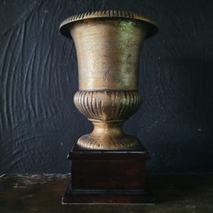 May include: A gold-painted urn with a fluted base sits on a dark wood pedestal. The urn is a decorative piece with a classic design.