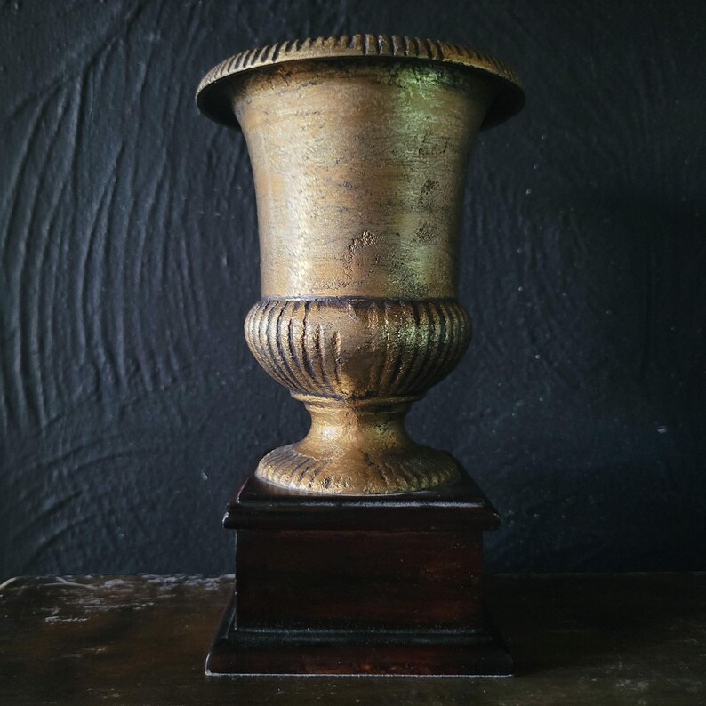 May include: A gold-painted urn with a fluted base sits on a dark wood pedestal. The urn is a decorative piece with a classic design.