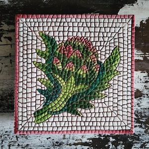 Puede incluir: Azulejo de mosaico cuadrado con diseño de alcachofa. La alcachofa se representa en tonos verdes y rosas, sobre un fondo de mosaico blanco y marrón con un borde rosa. Una pieza decorativa.