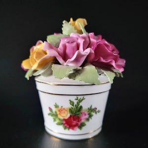 May include: A white porcelain pot with a gold rim, featuring a floral decal of red, yellow, and pink roses on the front. The pot is topped with a sculpted arrangement of three-dimensional ceramic roses in shades of pink, yellow, and green leaves.