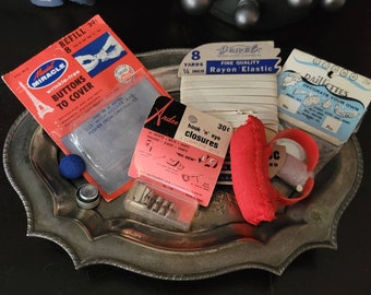 May include: A silver tray with various sewing and craft supplies, including a package of wrinkle-free buttons, a spool of white elastic, a package of hook and eye closures, a red foam hair roller, and a small blue button.