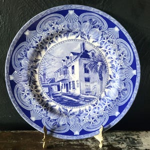 May include: A round ceramic plate featuring a blue and white transferware pattern. The center depicts a multi-story house with a balcony and palm tree, surrounded by a rope border. The rim displays a decorative motif of seashells and citrus fruit.