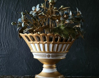 May include: A white porcelain vase with gold accents and a basket-like top, holding a bouquet of blue and green artificial flowers.