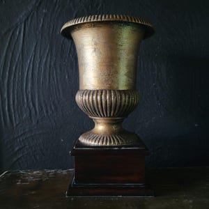 May include: A gold-toned metal urn with a fluted base sits on a dark wood base. The urn is a decorative piece for a home or office.