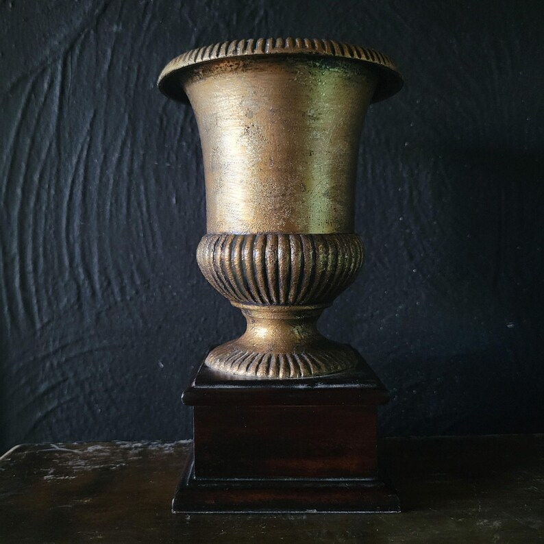 May include: A gold-toned metal urn with a fluted base sits on a dark wood base. The urn is a decorative piece for a home or office.