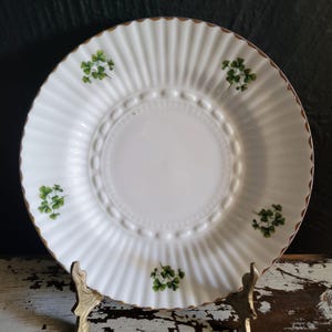 Puede incluir: Plato de cerámica blanca con diseño acanalado y borde dorado. El plato presenta grupos de tréboles verdes espaciados uniformemente alrededor del borde. Una pieza decorativa para servir o exhibir.